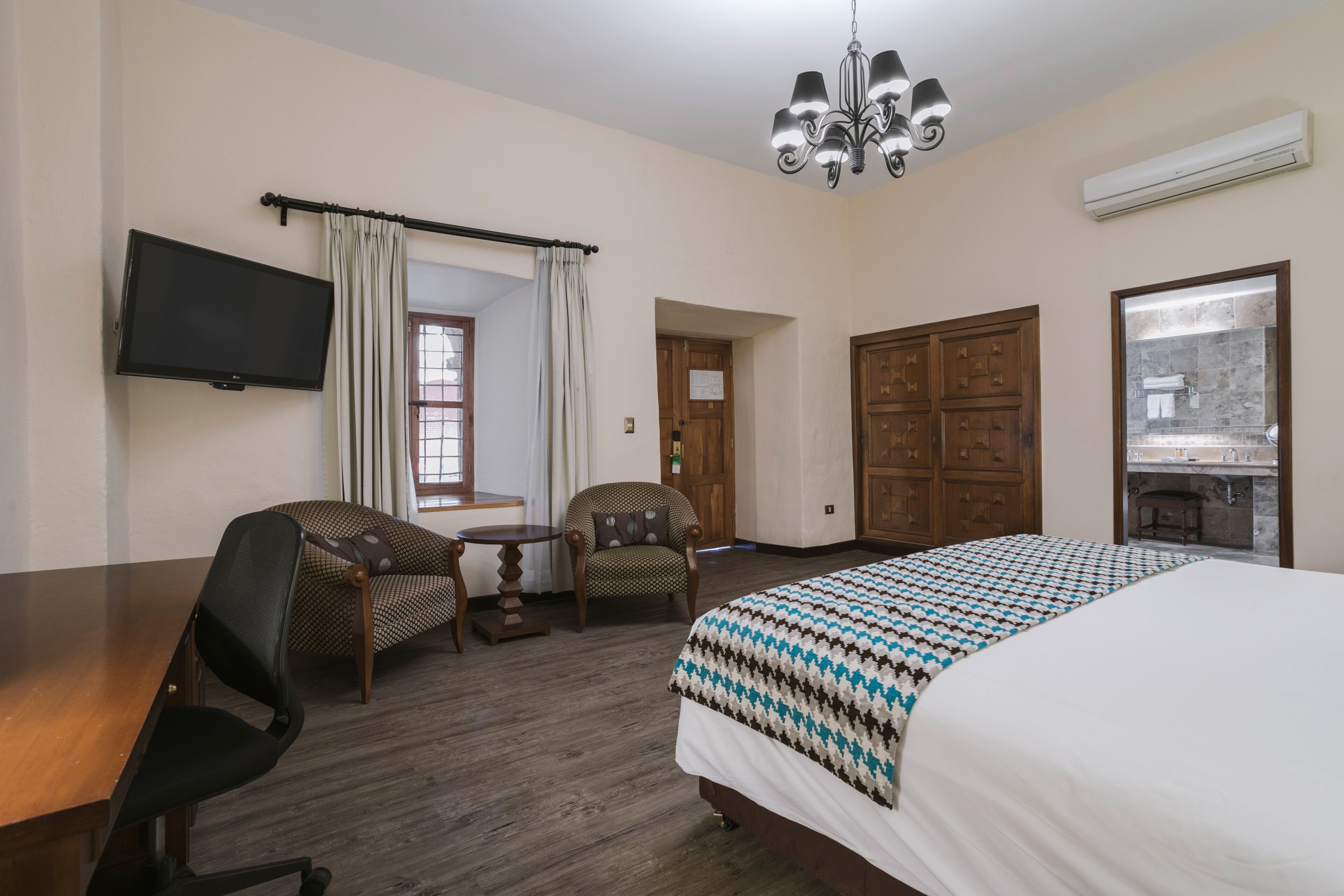 A hotel room with a large bed, two chairs, a small table, a TV, a chandelier, and a bathroom visible through a glass door.