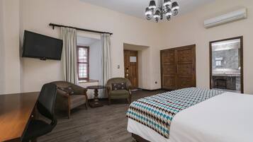 A hotel room with a large bed, two chairs, a small table, a TV, a chandelier, and a bathroom visible through a glass door.