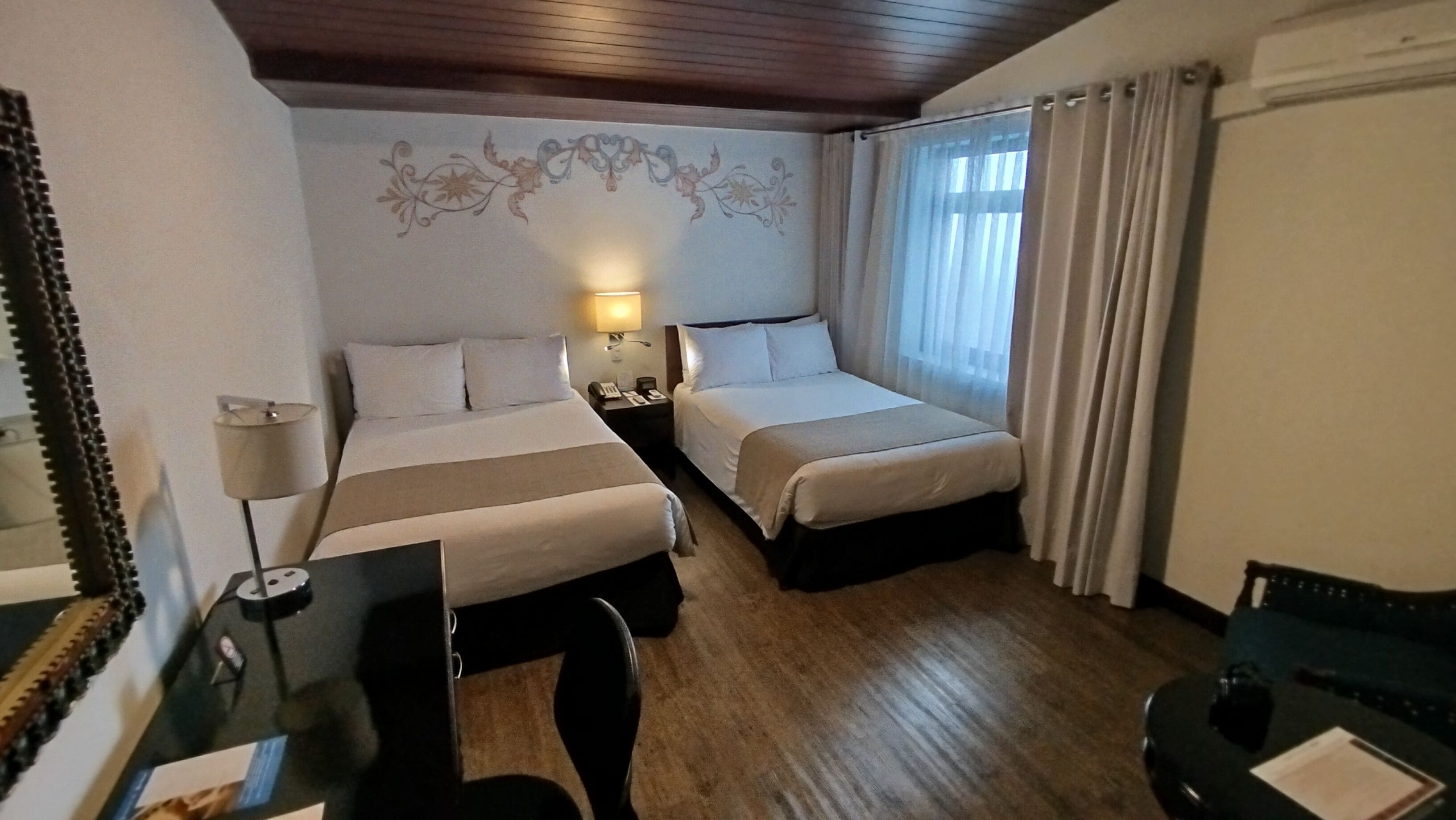 A hotel room with two beds, a desk, a chair, a lamp, and a mirror.