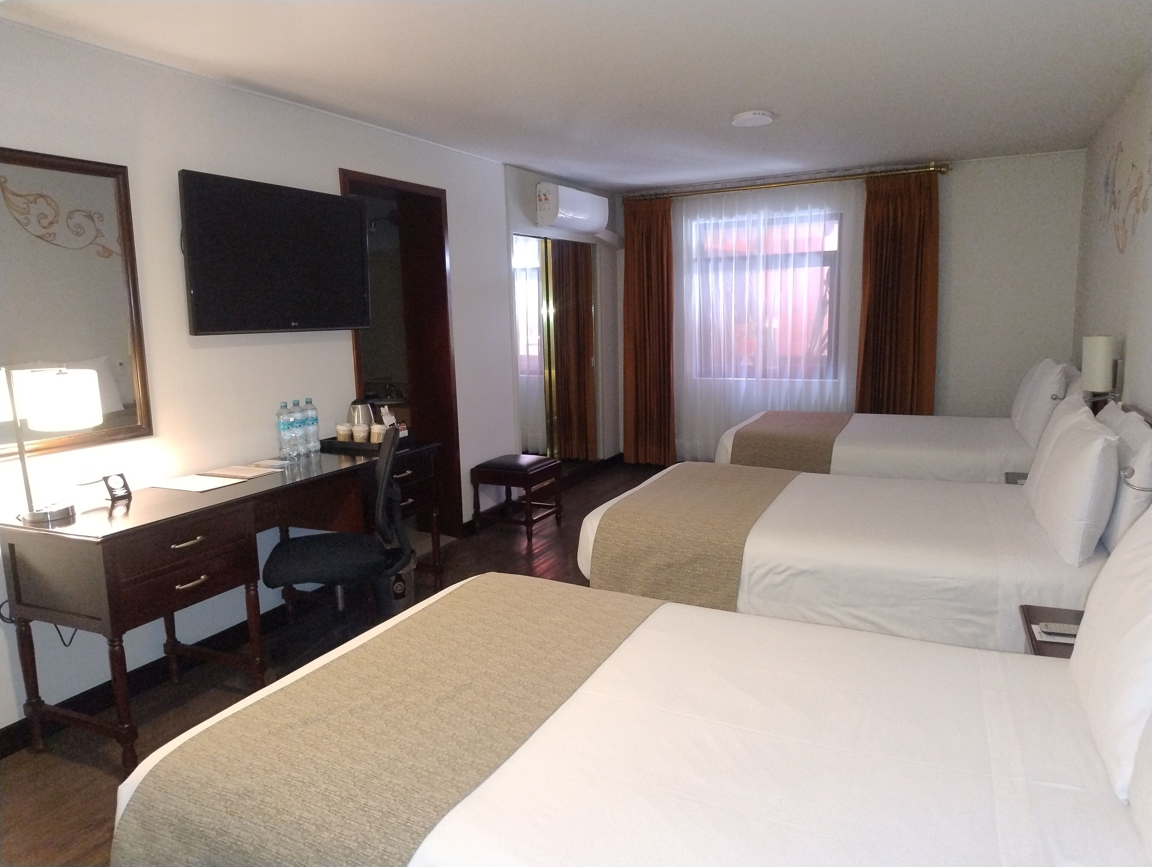 Ett hotellrum med två sängar, ett skrivbord, en stol, en spegel, en tv och ett fönster med gardiner.