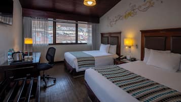 A hotel room with two beds, a desk with a chair, a lamp, and a view of the city through the window.
