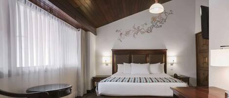 Habitación de hotel con techo de madera, una cama grande con ropa blanca, una mesita redonda y una silla.