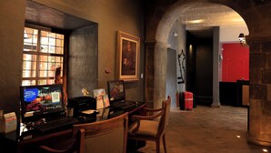 Rebedor d'hotel amb taulell de recepció, un ordinador i una cadira. Hi ha una finestra amb vista a l'exterior i una porta vermella al fons.