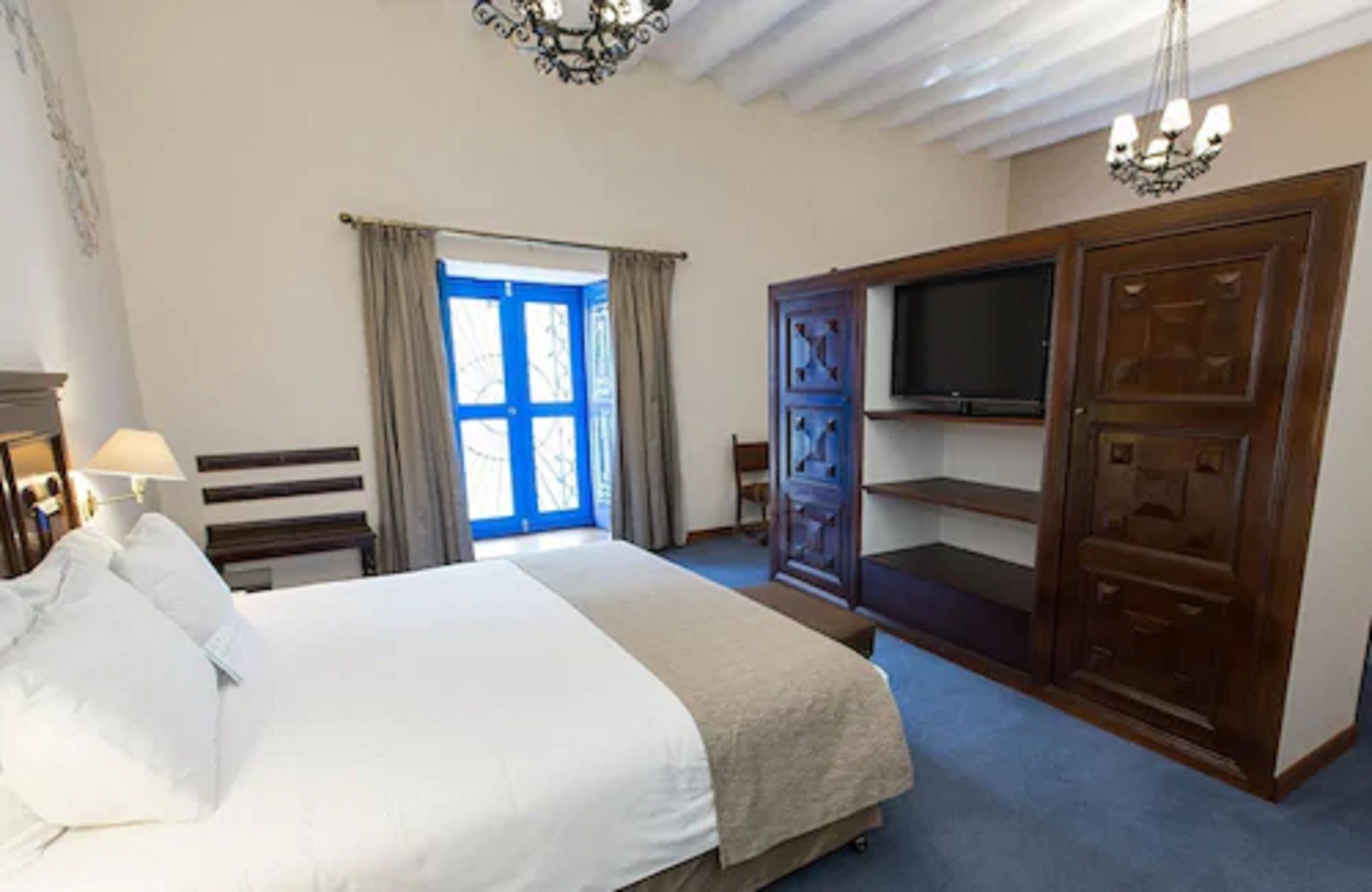 Une chambre à coucher avec un lit, un téléviseur posé sur un meuble en bois, une fenêtre avec des rideaux et un lustre.