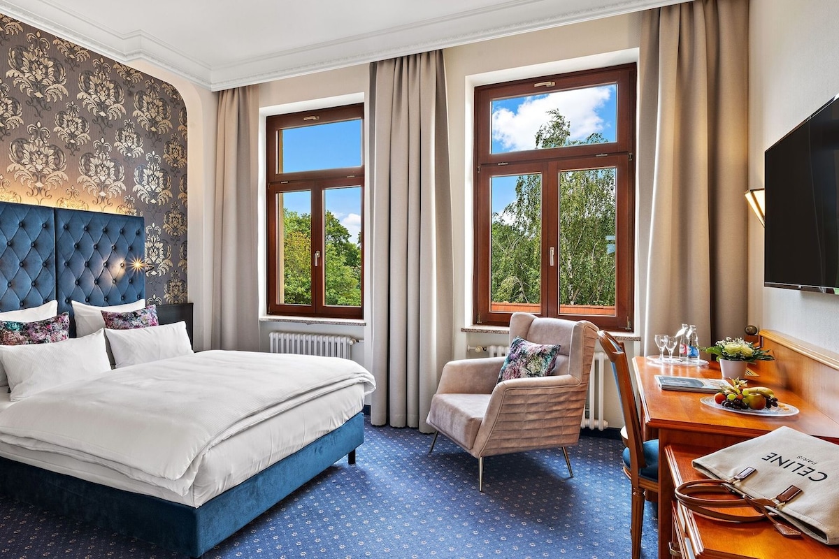 A hotel room with a large bed, a sitting area with a chair, a desk with a lamp, and a view of trees through the windows.