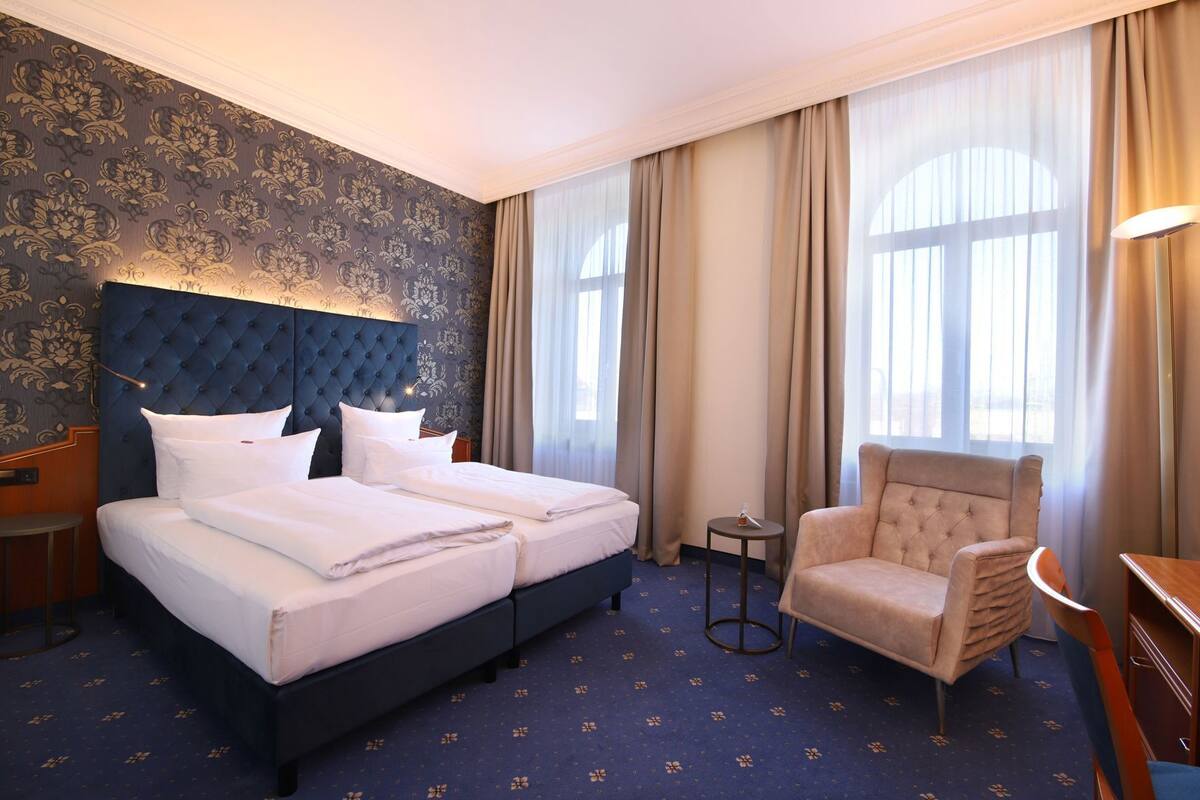 Une chambre d’hôtel avec un grand lit, une chaise, une petite table et une fenêtre avec des rideaux.