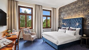 Habitación de hotel con una cama grande, un escritorio con silla y dos ventanas con vista a árboles.
