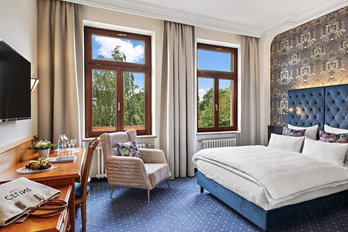 Une chambre d’hôtel avec un grand lit, un bureau avec une chaise et deux fenêtres donnant sur des arbres.