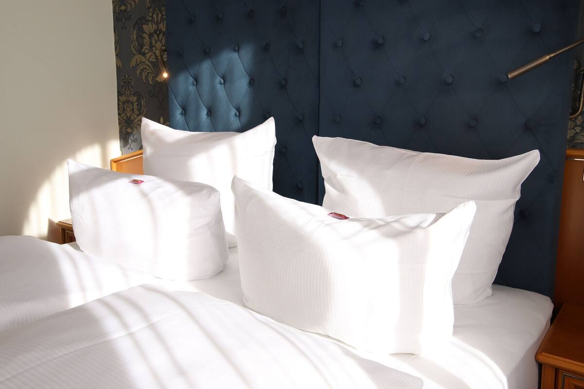 Un lit bien fait, avec du linge de lit blanc et des oreillers, une tête de lit capitonnée et une table de chevet en bois.