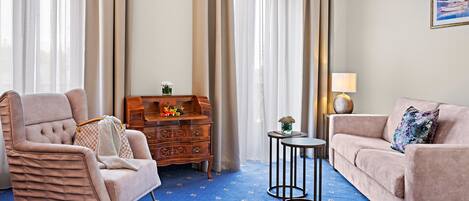 Habitación de hotel con alfombra azul estampada, cómoda de madera, sillón color beige, sofá y una mesita con lámpara.