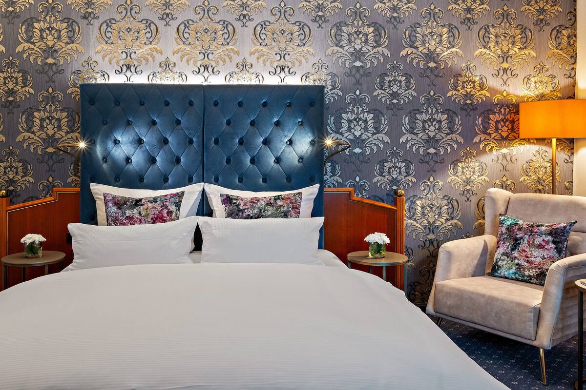 Une chambre d’hôtel avec un grand lit, une tête de lit capitonnée, deux tables de chevet, une chaise et un papier peint décoratif.
