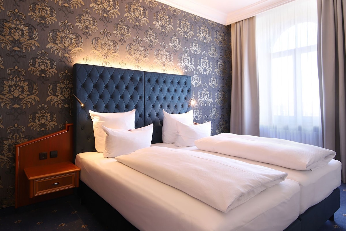 A hotel room with a tufted headboard, a wooden nightstand, a large bed with white linens, and a patterned wallpaper.