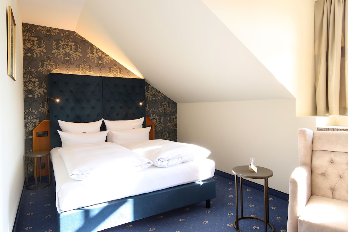A neatly made bed with a dark headboard, a small round table with a lamp, and a plush armchair in a hotel room with patterned wallpaper and a sloped ceiling.
