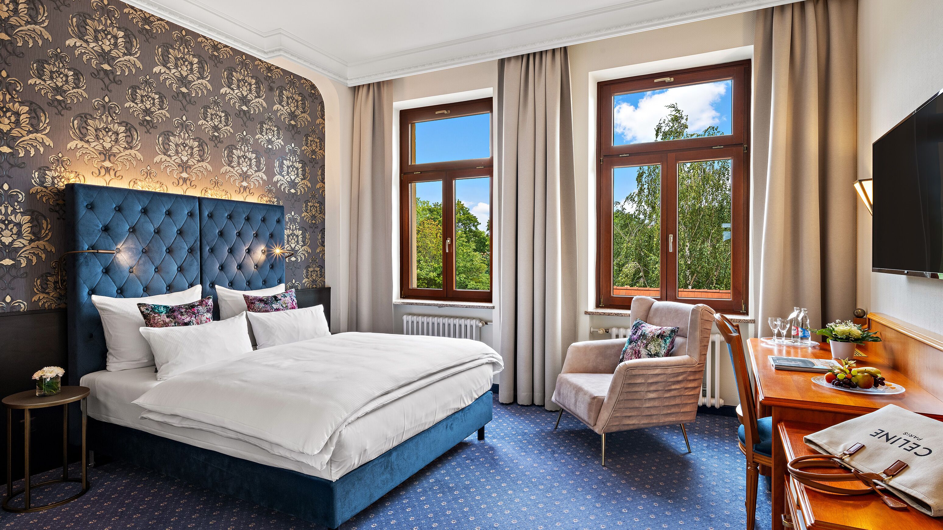 Una camera d'albergo con un letto grande, una poltrona, una scrivania con una lampada e due finestre con tende.