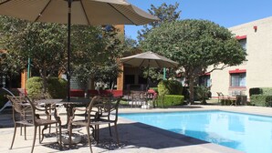 A poolside area with a table and chairs, surrounded by trees and a building in the background.