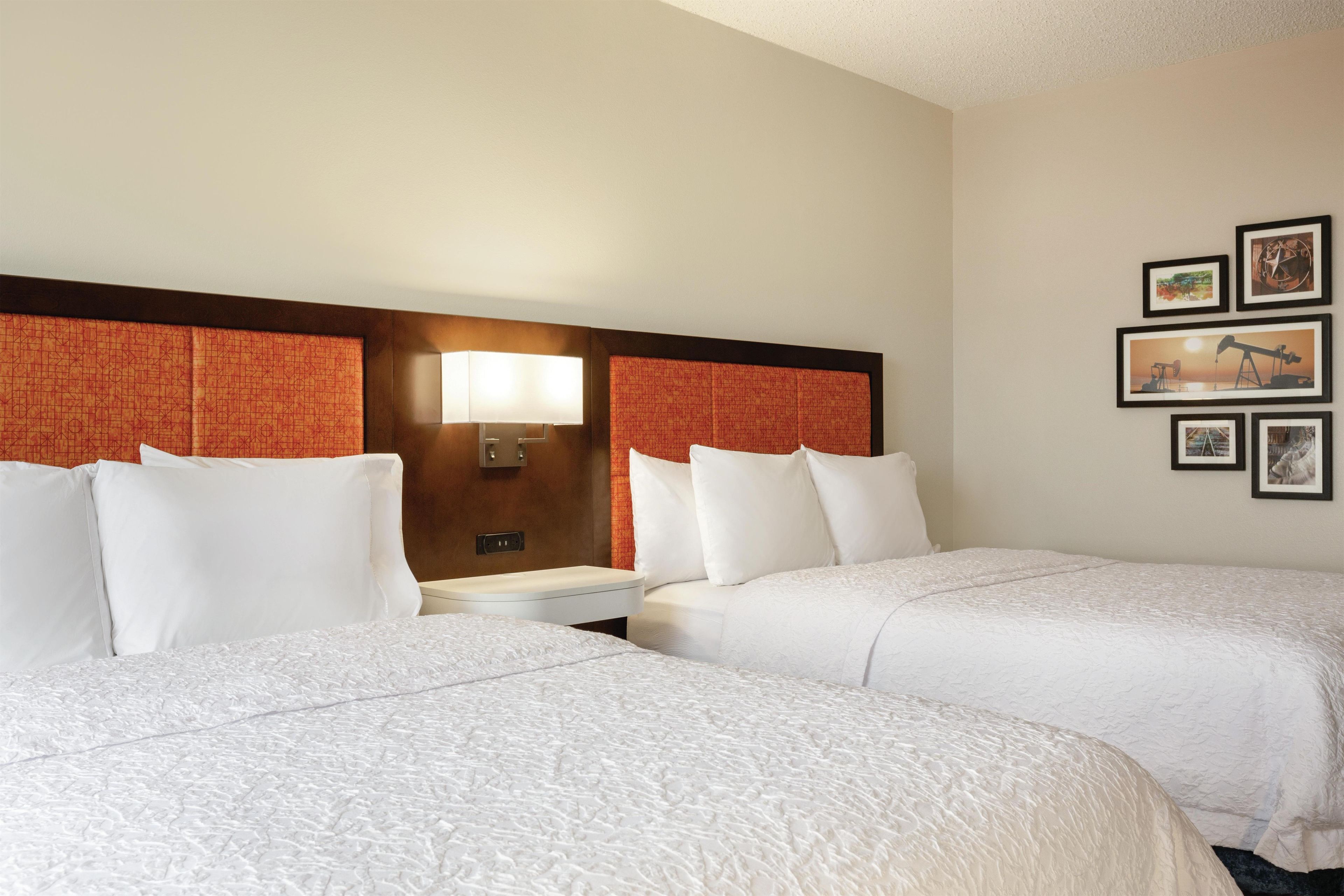 A hotel room with two beds, a nightstand, a wall-mounted lamp, and framed pictures.