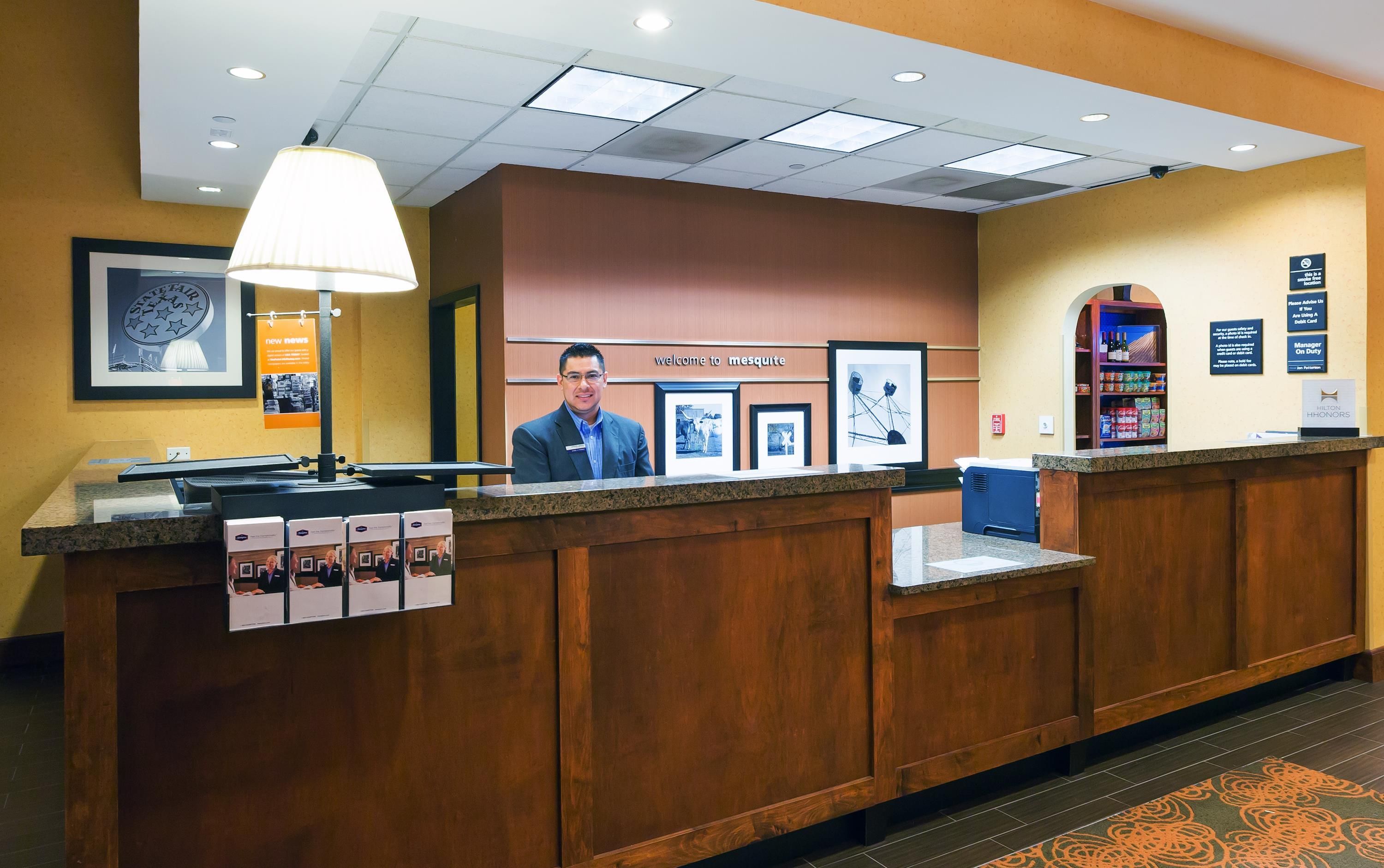 Una recepción de hotel con un cartel de bienvenida enmarcado, una foto enmarcada de una persona, una lámpara y un minibar.