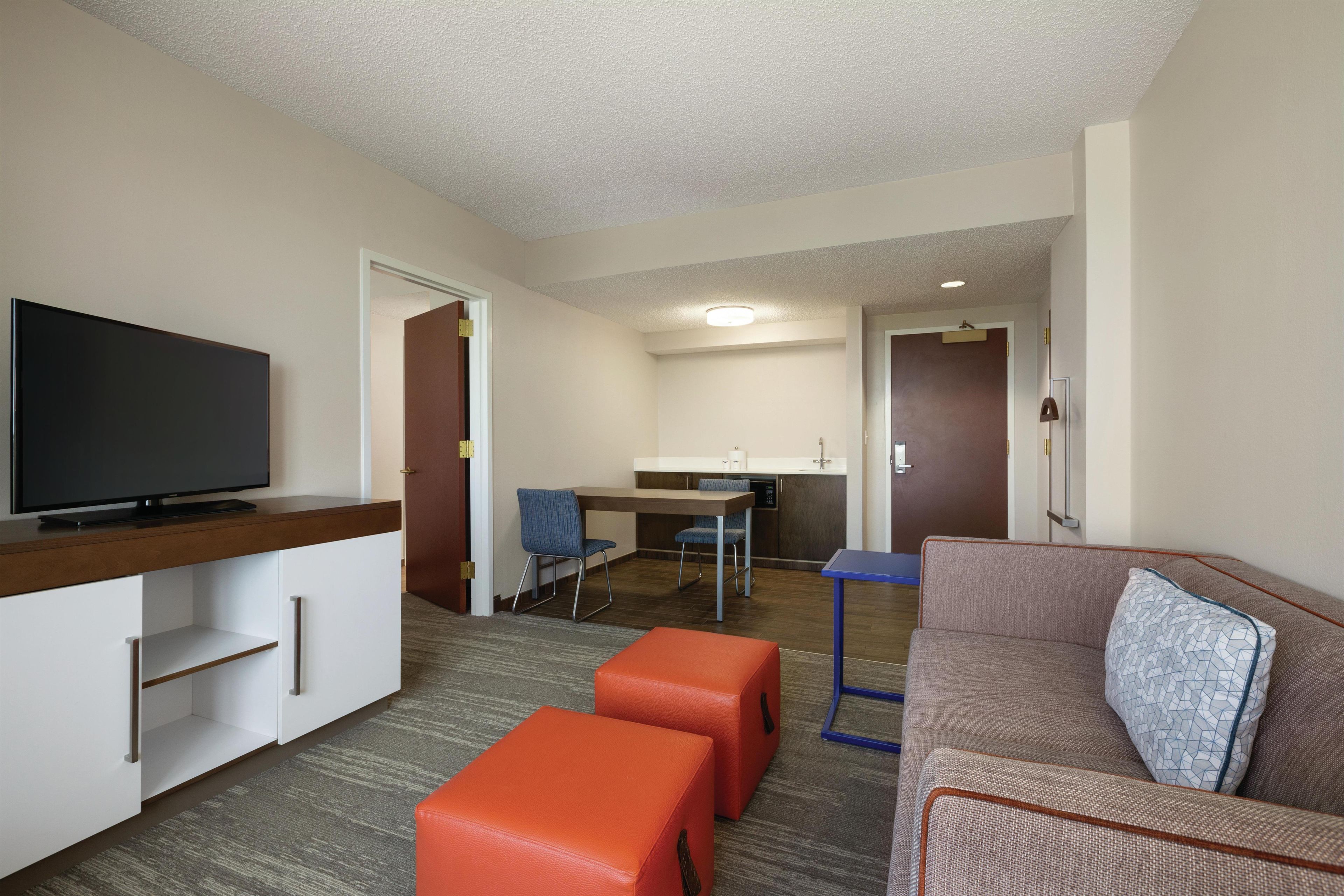 Habitación de hotel con televisión, kitchenette, comedor y zona de estar con sofá y otomanas.