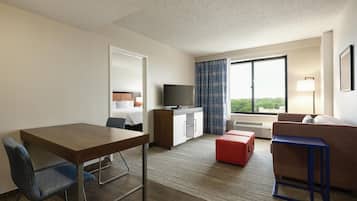 A hotel room with a wooden desk, a blue chair, a red ottoman, a sofa, a television, a window with curtains, and a bed with a headboard.