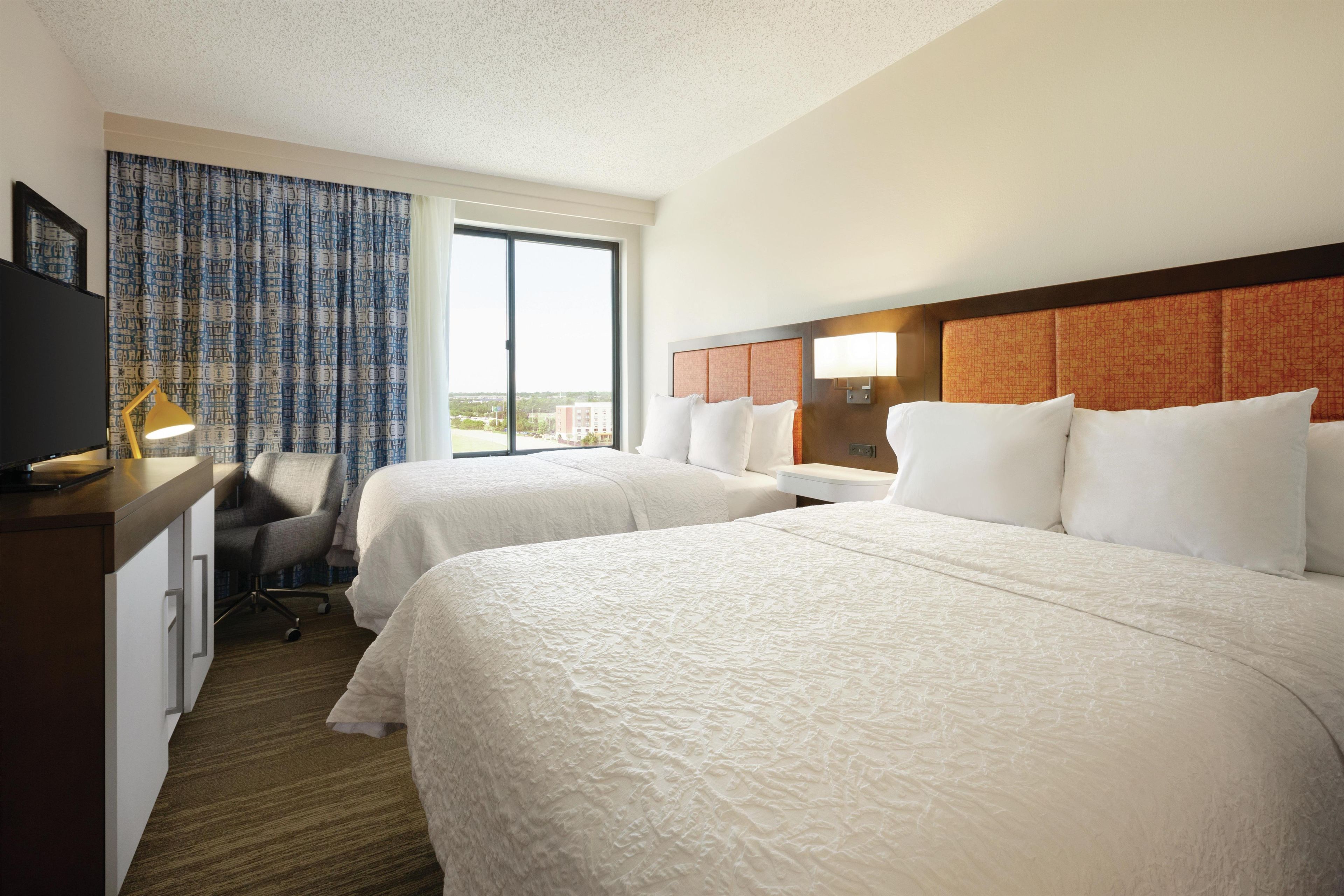 A hotel room with two beds, a desk with a lamp, a chair, and a large window.