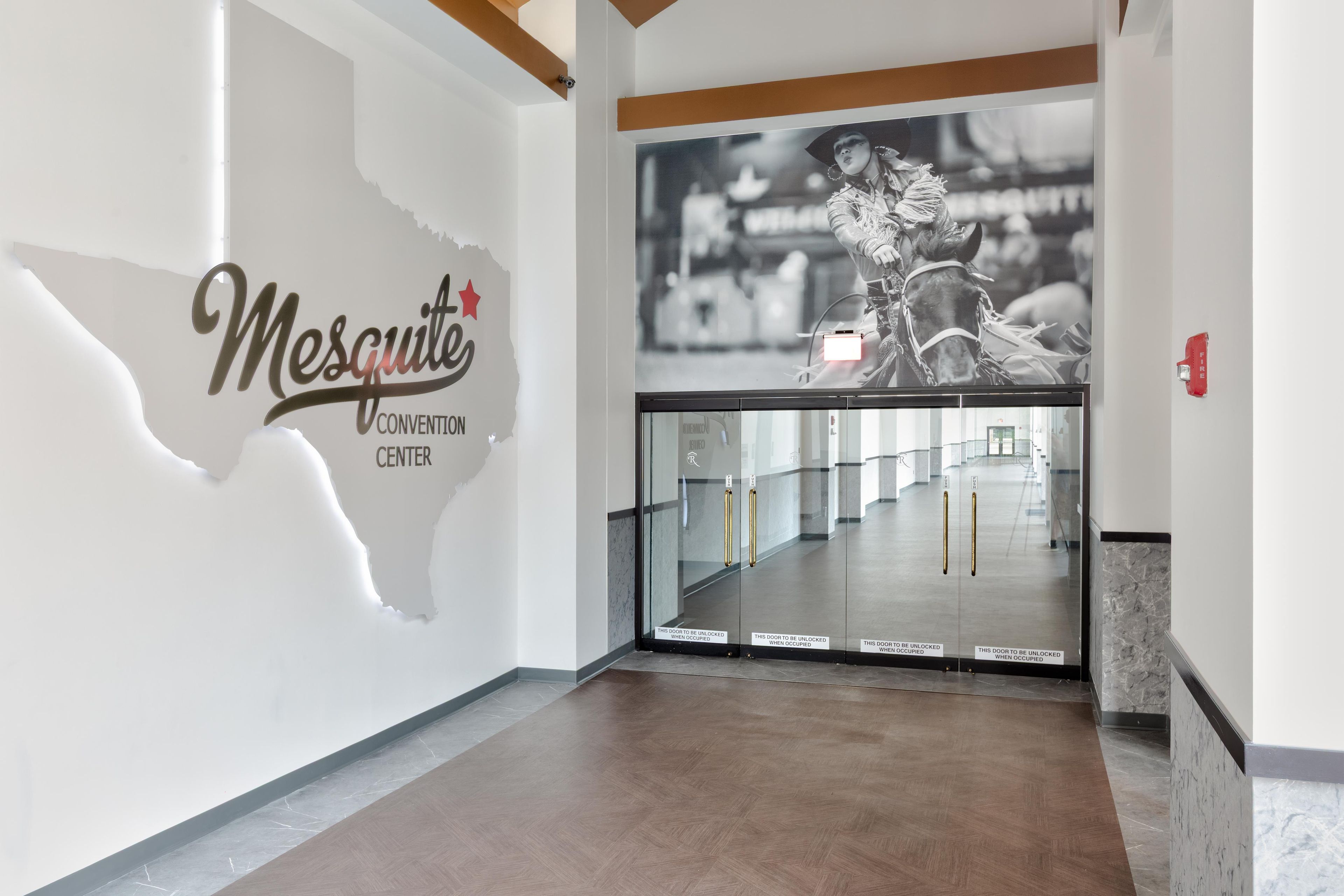 A hallway with a glass door leading to a room, a wall with the text 'Mesquite Convention Center,' and a poster of a person riding a horse.
