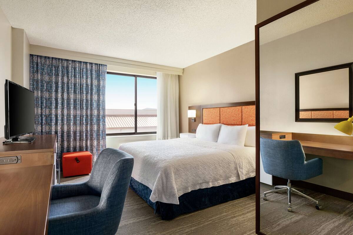 Habitación de hotel con cama, escritorio, silla, televisión y ventana con cortinas.