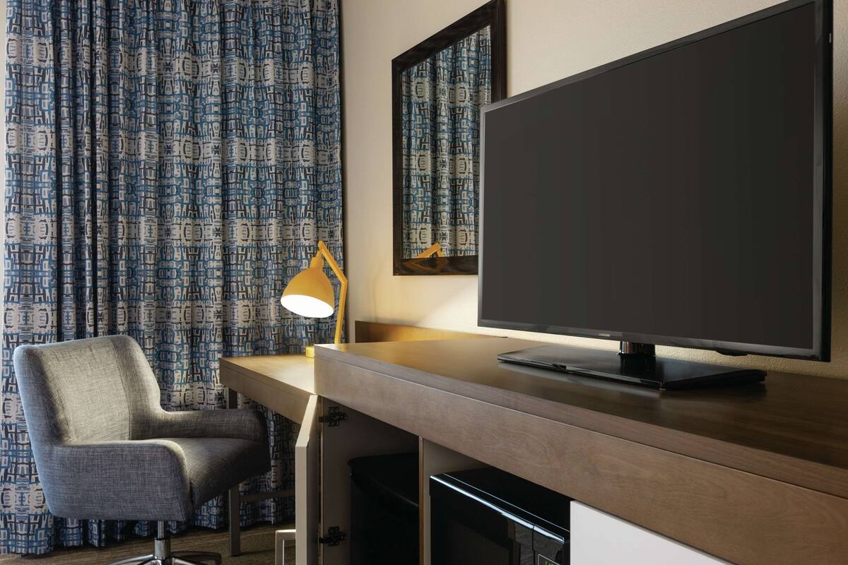 Una habitación de hotel con un escritorio de madera, un televisor de pantalla plana, una silla y cortinas con estampado.
