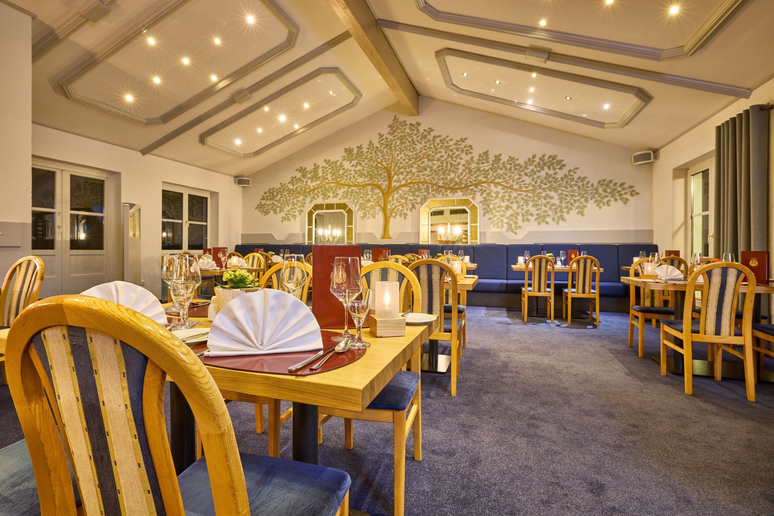 A dining area with wooden tables and chairs, a ceiling with recessed lighting, and a wall mural of a tree.
