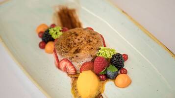 Un plato de postre con una porción de pastel, fresas, frambuesas, zarzamoras y una cucharada de salsa amarilla.