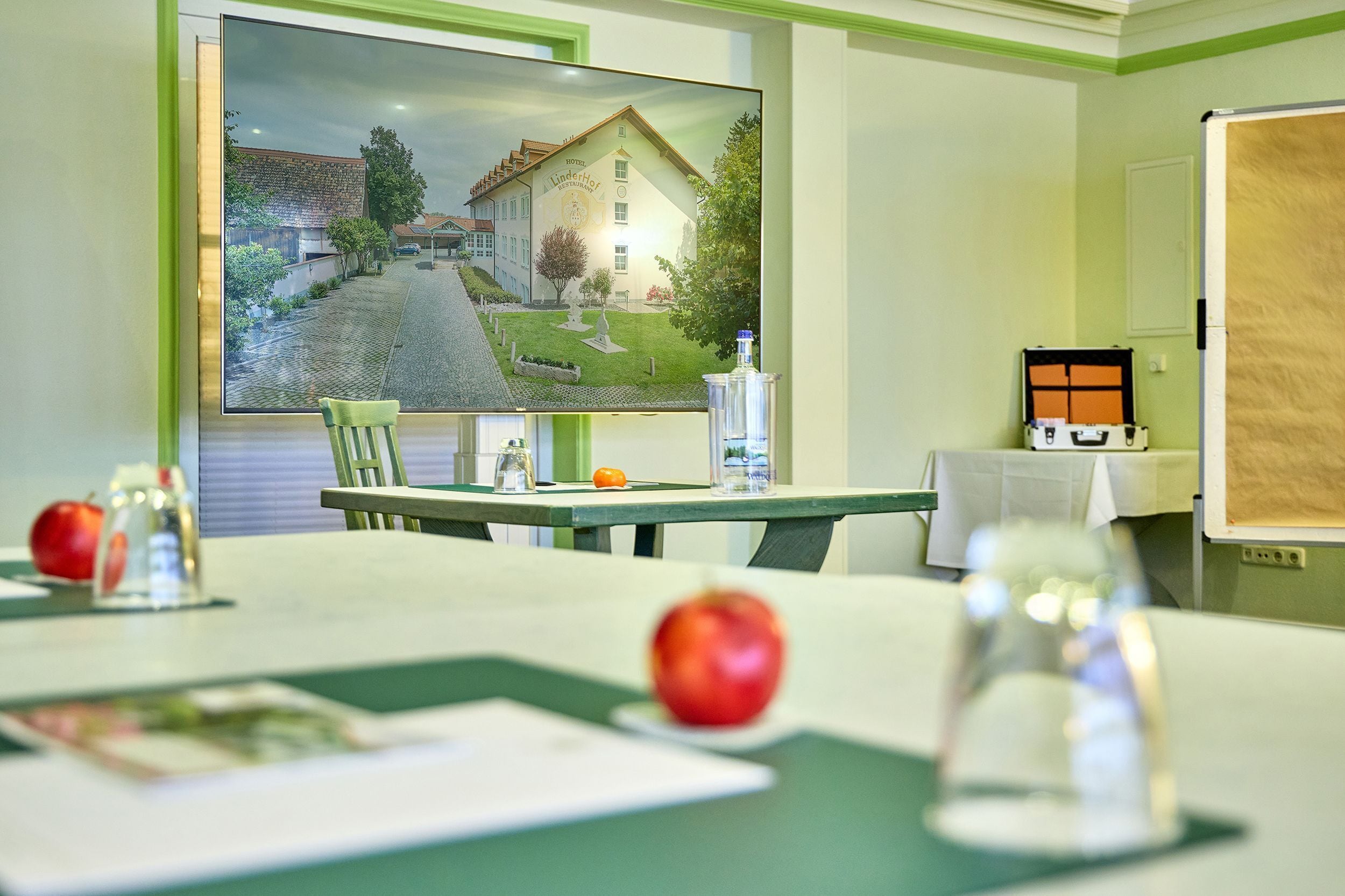 A room with a large flat-screen TV displaying a scenic view, a table with a green tablecloth, two apples, a glass, and a bottle.