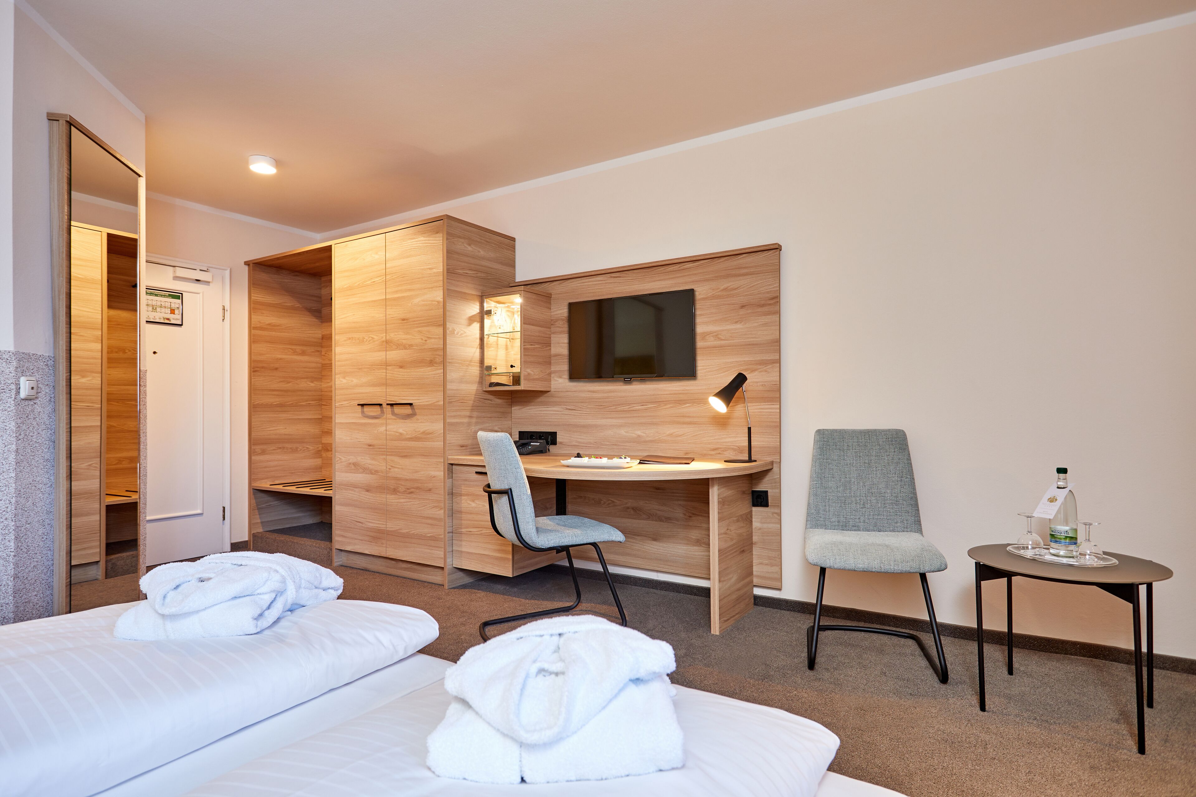 Quarto de hotel com duas camas, uma escrivaninha, uma cadeira, uma televisão e uma mesinha com garrafa e copos.