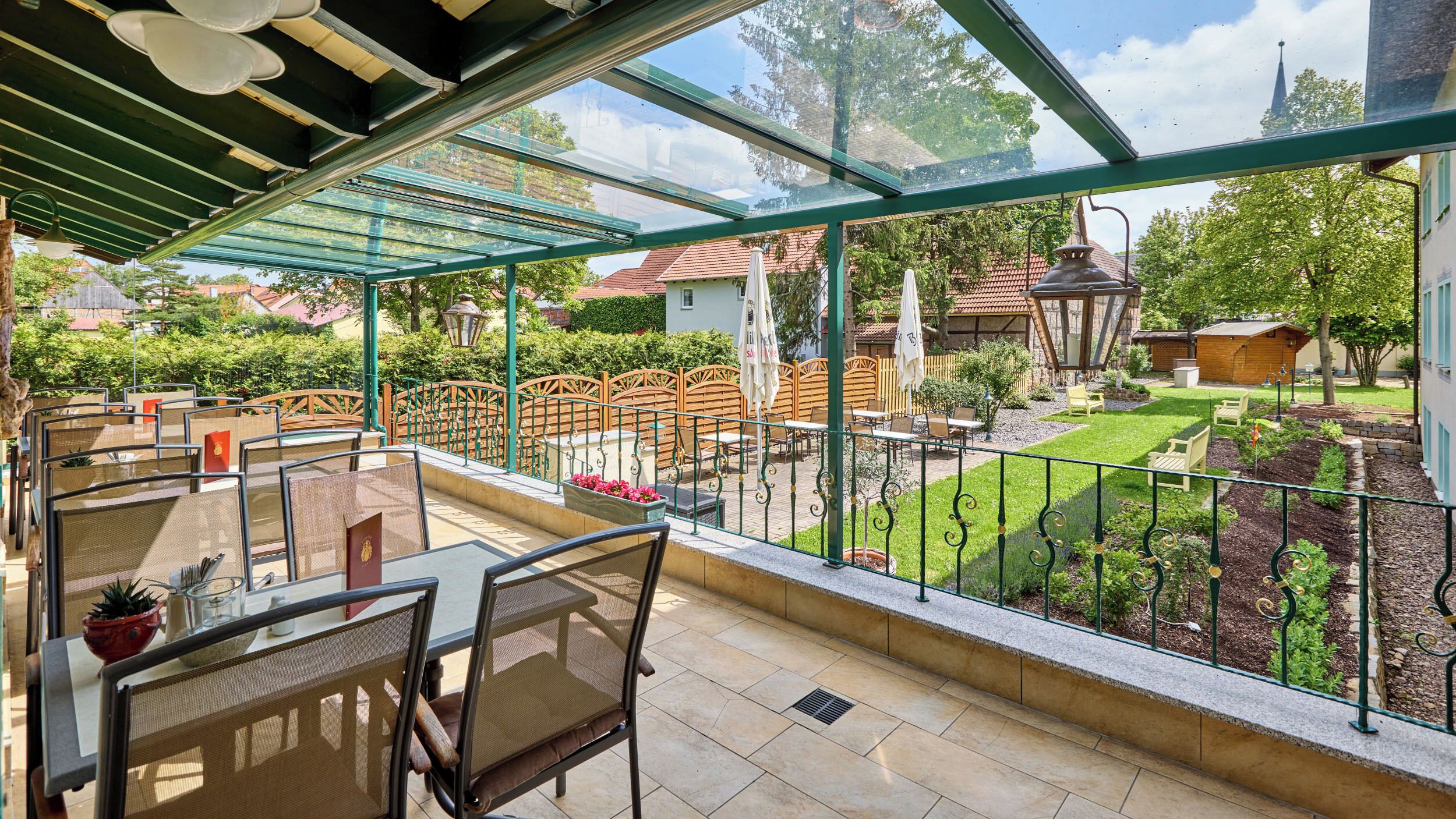 A covered outdoor patio with tables and chairs, a view of a garden, and a building in the background.