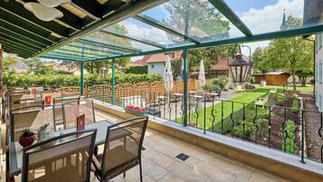 A covered outdoor patio with tables and chairs, a view of a garden, and a building in the background.