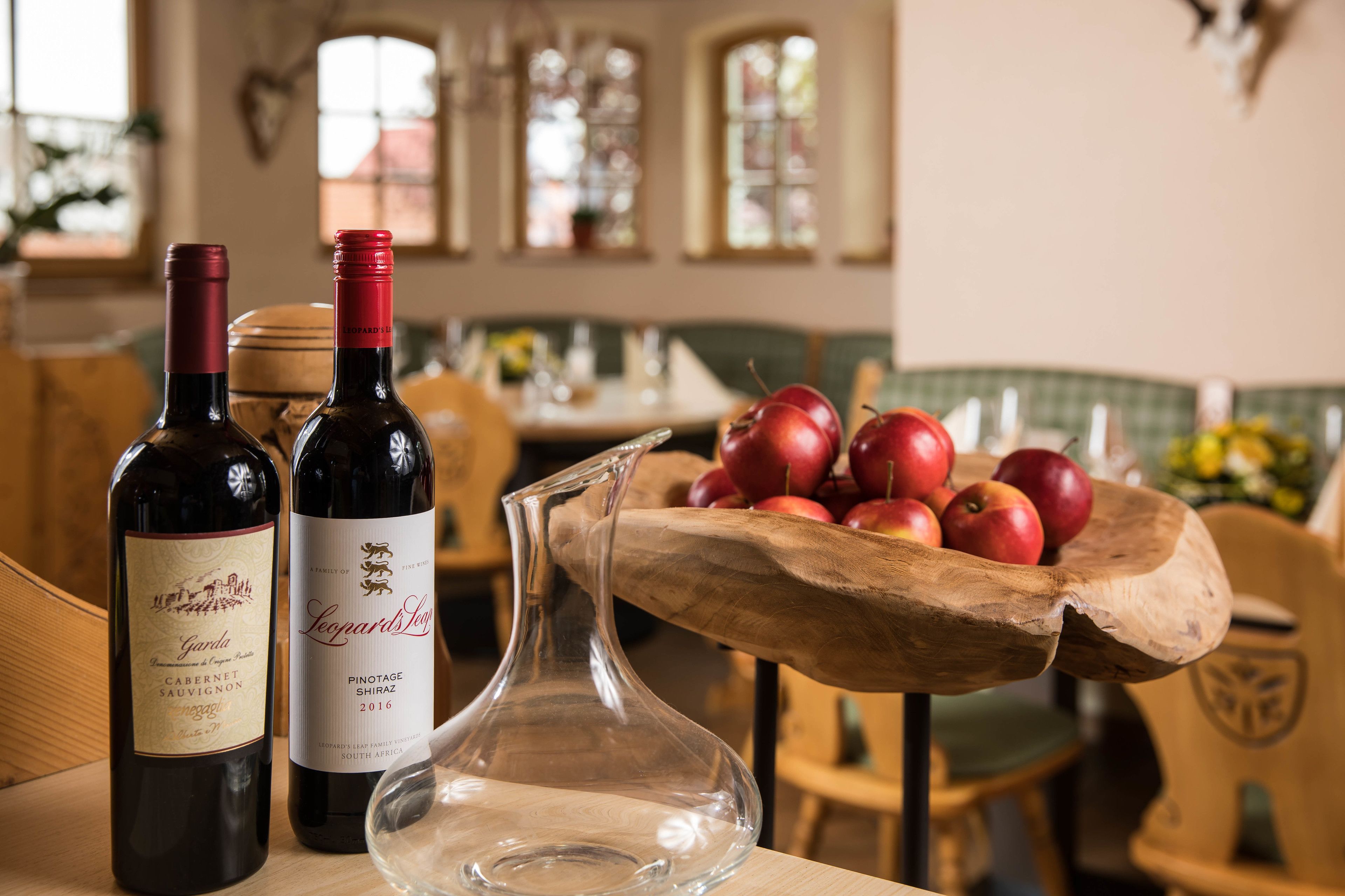 Two wine bottles and a pitcher on a table with a wooden bowl of apples in the background.