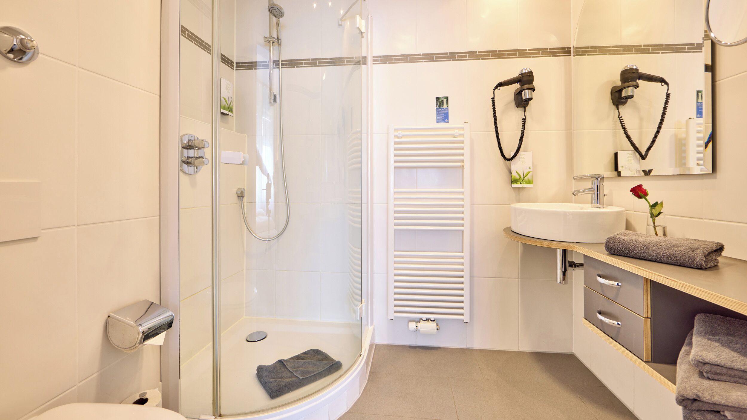 Un baño moderno con ducha de vidrio, lavamanos blanco y tocador con toallero.