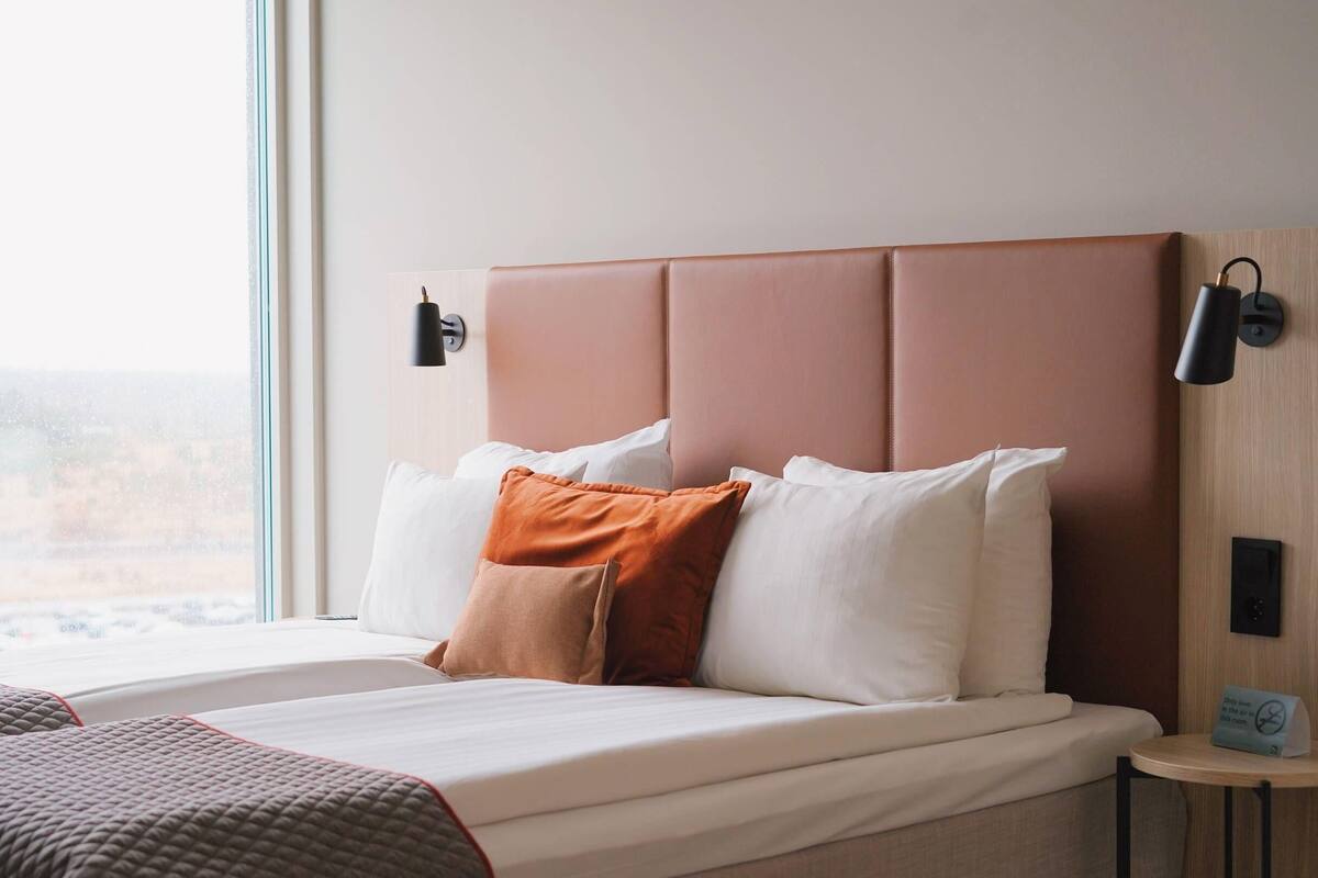 Een hotelkamer met een bed, kussens, een nachtkastje en een raam met uitzicht.