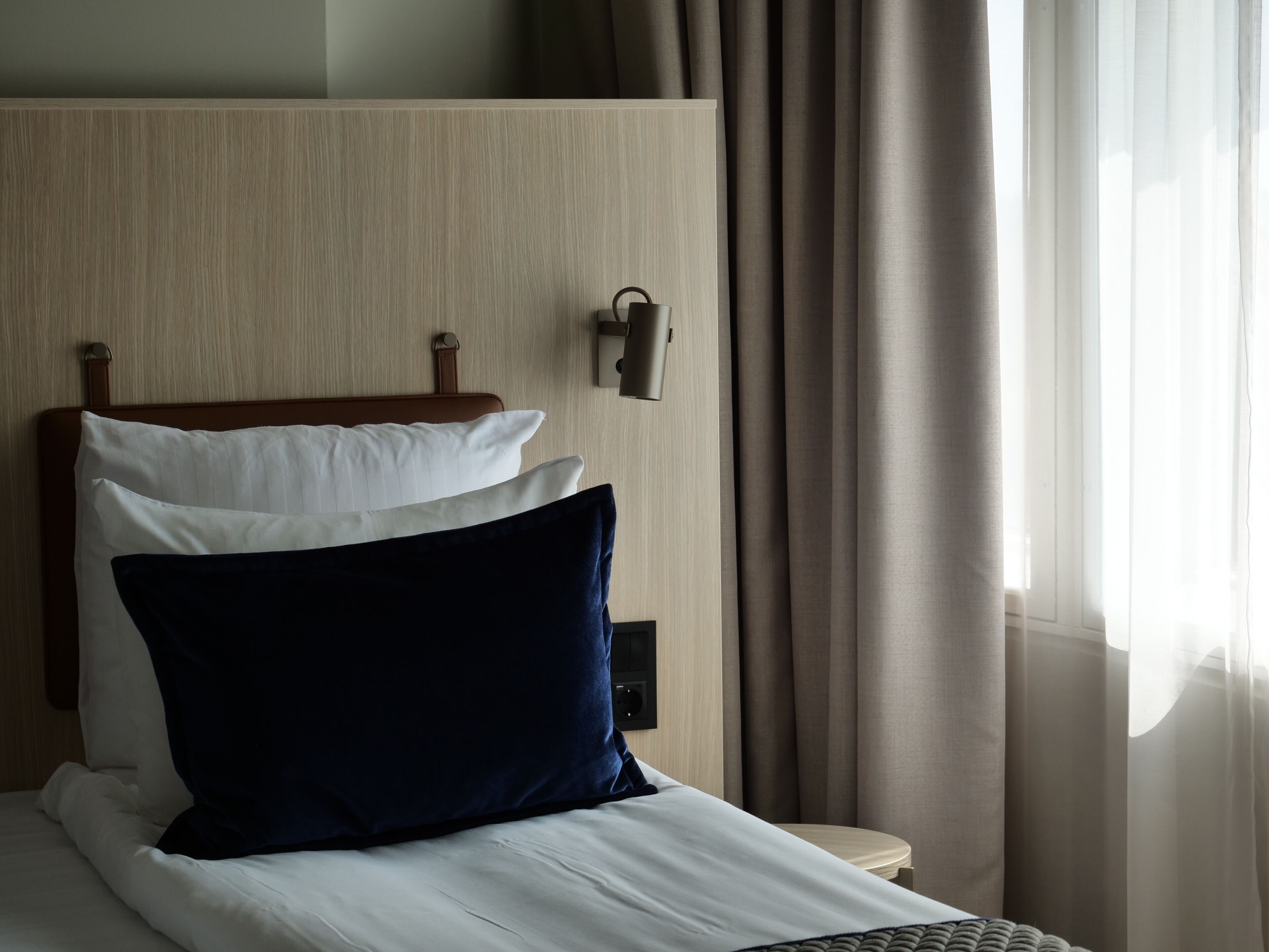 Una cama bien hecha con almohadas blancas y azul oscuro, un cabecero de madera, una mesita de noche y una lámpara fijada a la pared.