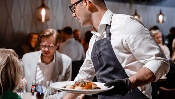 Un serveur apporte des plats dans un restaurant au décor moderne, avec d’autres clients assis à des tables à l’arrière-plan.