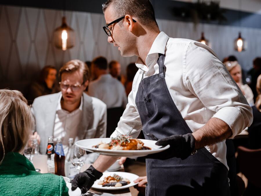 Un serveur apporte des plats dans un restaurant au décor moderne, avec d’autres clients assis à des tables à l’arrière-plan.