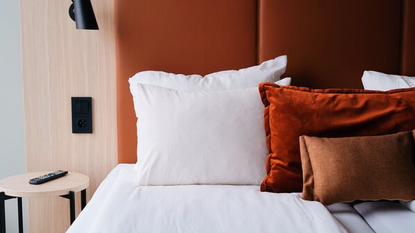 A neatly made bed with white and brown pillows, a remote control on a small table beside it, and a wall-mounted light fixture.