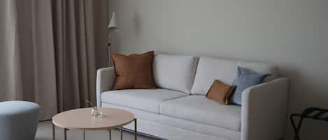 A modern living room with a light grey sofa, two cushions, a round wooden coffee table, a grey ottoman, and a floor lamp.