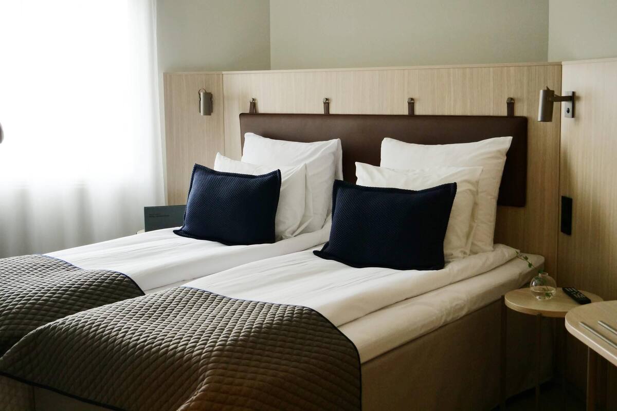 Een hotelkamer met twee bedden, elk met hoofdbord, wit beddengoed en donkerblauwe kussens. Er staat een klein nachtkastje met een lamp en een afstandsbediening.