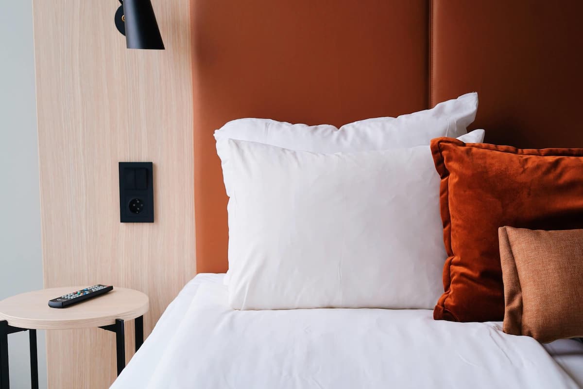 Een net opgemaakt bed met witte en bruine kussens, een afstandsbediening op een klein tafeltje ernaast, en een zwart wandpaneel.