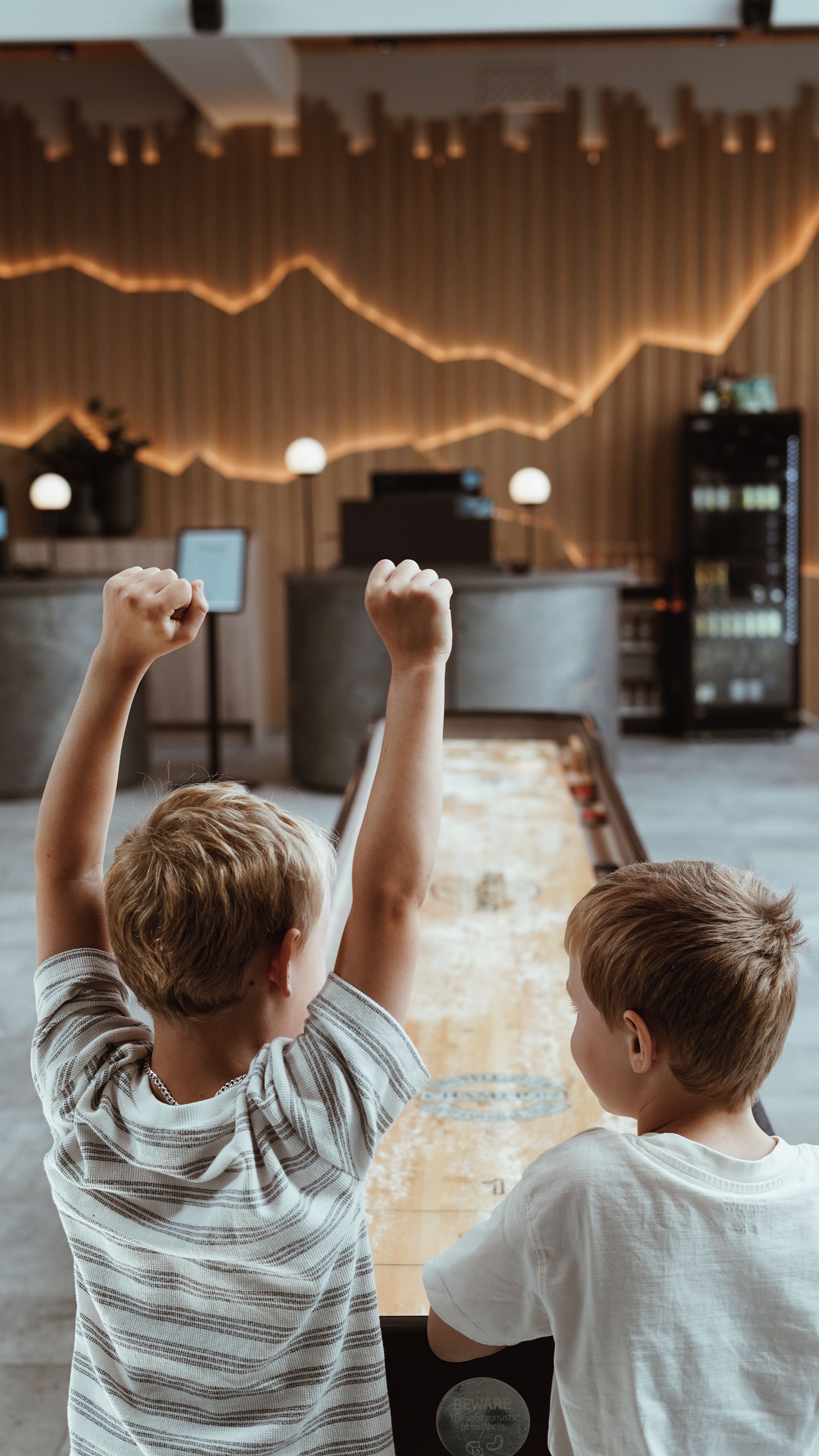 Two children raising their hands in a room with a modern interior design, including a wooden wall and a reception desk.