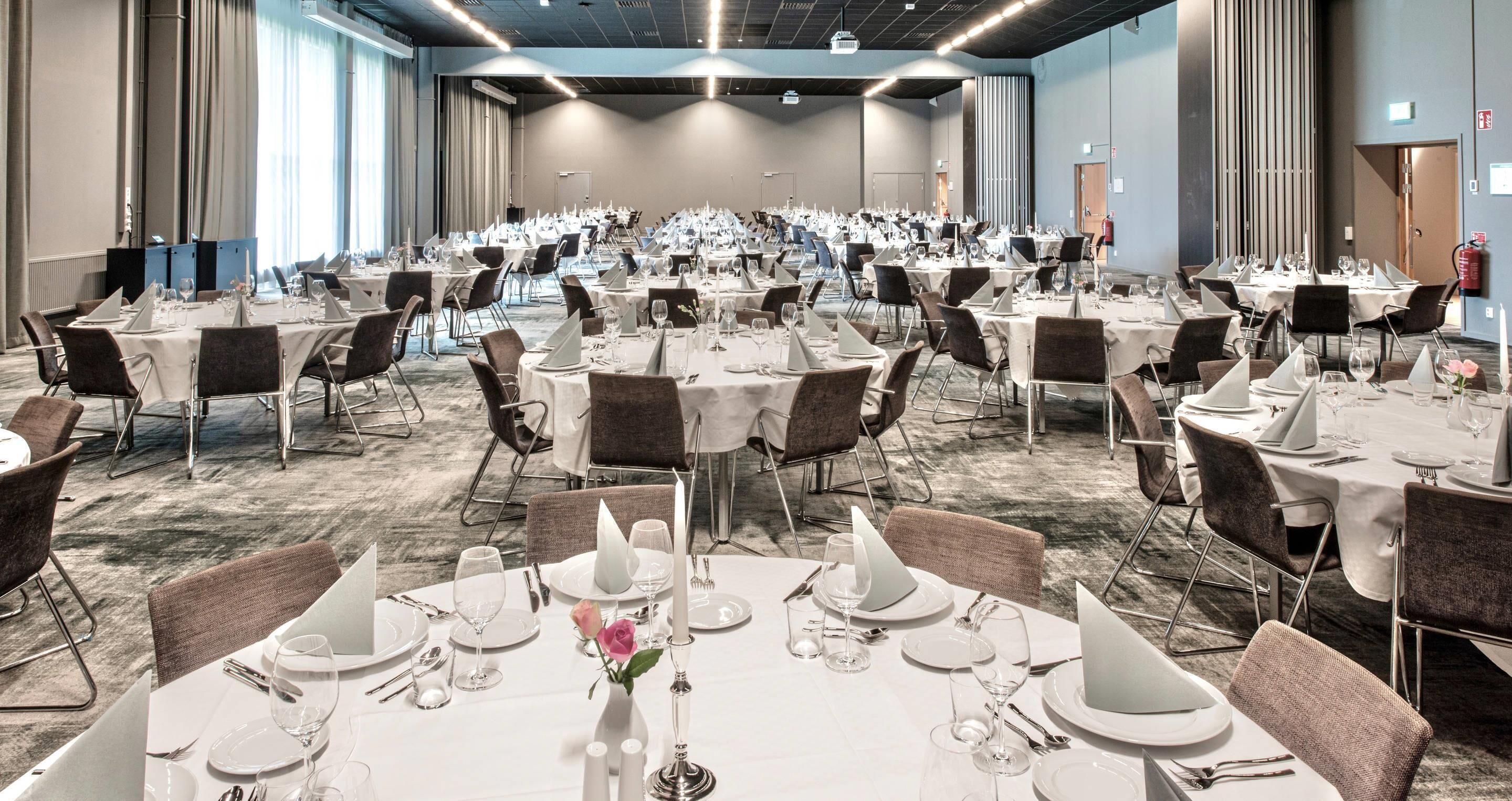 Een ruime balzaal met meerdere ronde tafels gedekt voor een diner, elk met witte tafelkleden, borden en bestek.