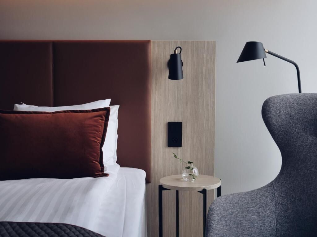 Una camera d'albergo moderna con un grande letto, un comodino con un vaso, una lampada e una poltrona grigia.