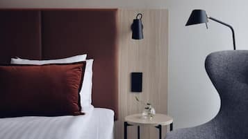A modern hotel room with a large bed, a side table with a vase, a lamp, and a grey armchair.