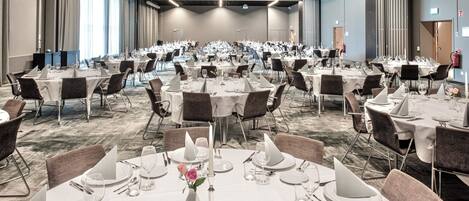 A large banquet hall with round tables set for dining, featuring white tablecloths, silverware, and glassware.
