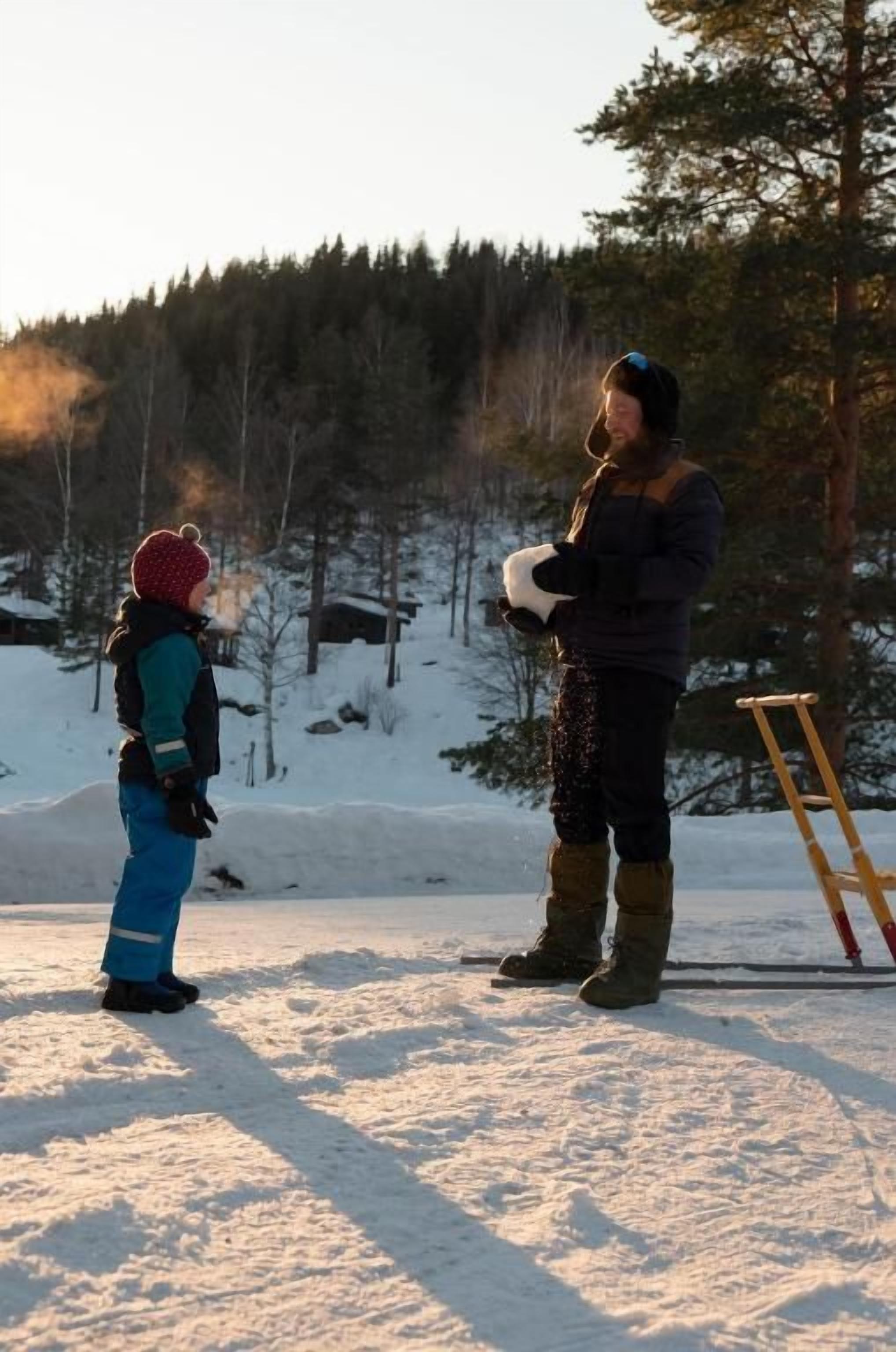 A snowy outdoor scene with a person standing and holding a snowboard, another person standing nearby, and a wooden ladder leaning against a tree.
