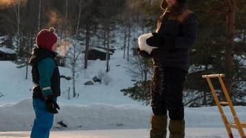 A snowy outdoor scene with a person standing and holding a snowboard, another person standing nearby, and a wooden ladder leaning against a tree.